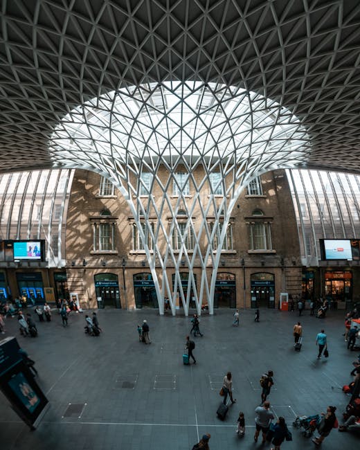 Inside a large, modern train station with a high, geometric ceiling constructed from a network of intersecting metal beams forming triangular patterns. Natural light filters through the ceiling, illuminating the spacious concourse below. The area features a mix of historic brickwork and contemporary glass walls, with multiple digital screens displaying travel information and advertisements. Numerous passengers are scattered across the open floor, some walking with luggage, others waiting or standing in small groups. Several people are seen carrying boxes, bags, or small furniture items as part of a home relocation or moving process, facilitated by a professional removals service such as Man and Van King's Cross. The scene depicts the loading or unloading phase of a furniture transport effort, with individuals assisting in carrying items towards or from waiting vehicles, highlighting efficient logistics involved in moving within or through this busy transport hub.