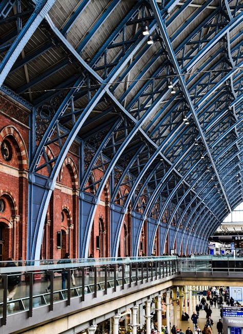 A wide view of King's Cross train station with a high, curved steel and glass roof supported by blue-painted metal arches, spanning over a spacious interior with red-brick walls featuring arched windows. The station’s interior includes multiple levels, with a balcony and glass safety barriers overlooking the busy lower level where numerous people are walking and waiting. Visible are large digital departure screens, signage, and part of a platform area in the background. The scene is well-lit by natural light filtering through the glass roof, highlighting the architectural details of the station. This setting depicts a typical scene during a home relocation or moving process, with the station's large open space suitable for the transportation and loading of furniture and boxes, often facilitated by professional removals services such as those offered by Man and Van King's Cross.