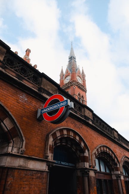 Photograph showing the exterior of a brick building with a prominent London Underground roundel sign attached to it, indicating an underground station entrance beneath three arched windows. Above the building, part of a historic clock tower with a tall, pointed spire is visible against a partly cloudy sky. The scene captures the urban environment typical of King’s Cross, with the clock tower and underground station sign illustrating a common setting for house removals and furniture transport involving building access points and city infrastructure. The image emphasizes the typical exterior elements encountered during home relocation or moving services provided by companies like Man and Van King's Cross, highlighting aspects of moving logistics such as building entry and city infrastructure in Camden.