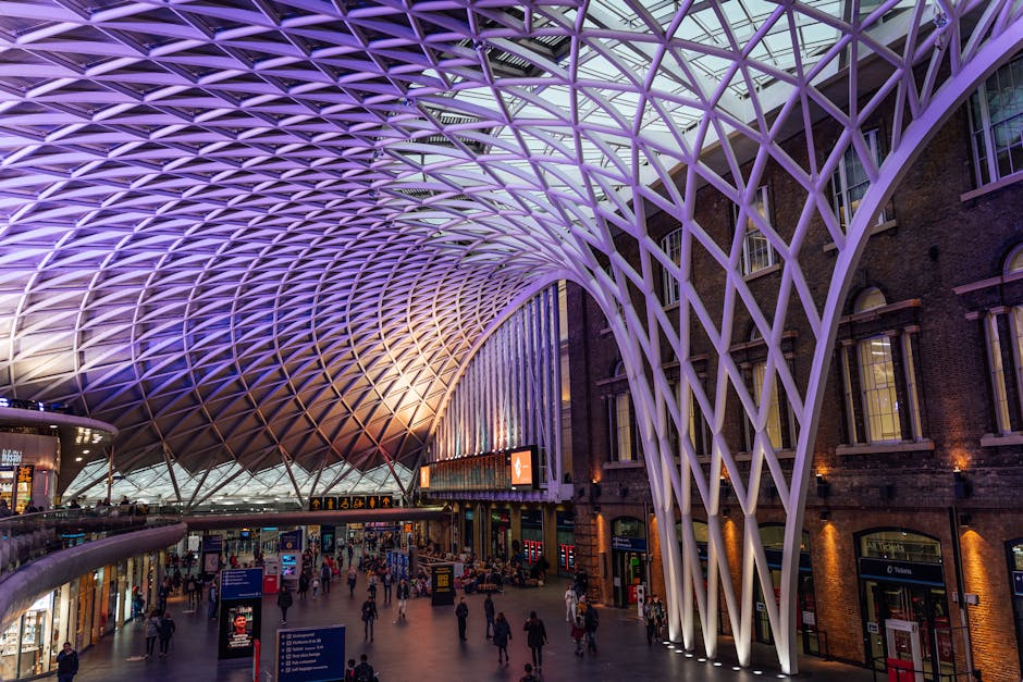 Inside King's Cross station, a large modern canopy with a geometric lattice pattern illuminated with purple and blue lighting spans the ceiling, casting a glow over the bustling interior filled with people walking and standing in the spacious open area. Below the canopy, various shops and signage are visible along the perimeter, with a mix of brick and glass buildings lining the station’s interior. Moving equipment such as hand trucks, cardboard boxes, and padding materials may be seen as part of home relocation or furniture transport preparations for house removals. The scene captures the busy atmosphere of a major transport hub, emphasizing the importance of careful packing, loading, and logistical planning during a home relocation process, which is relevant to services offered by Man and Van King's Cross that facilitate efficient packing and moving services.