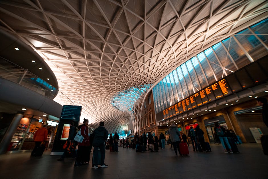 Inside the modern train station at Kings Cross, the spacious hall features a high, intricately patterned ceiling with a geometric lattice design made of metal or wood, illuminated by soft ambient lighting. Large glass windows along the upper walls allow natural light to fill the area, creating a bright and airy atmosphere. Several travelers are visible, some standing, others walking with rolling suitcases or carrying bags, and a few are engaged in consulting electronic departure boards that display train schedules in orange text. The station floor is tiled and uncluttered, with some luggage carts and small seating areas visible along the sides. In the background, retail outlets and cafes can be seen, with interior lighting contributing to the lively but orderly environment typical of a busy transportation hub. This scene exemplifies the dynamic space where passenger flow and luggage movement are central to the station's function, relevant to home relocation and furniture transport logistics managed by companies like Man and Van King's Cross, supporting efficient packing and loading processes for journeys or removals.