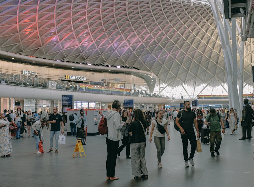 The spacious interior of a busy modern train station with a high, curved ceiling featuring a diamond-patterned lattice design illuminated by pink and purple lighting. The station is filled with numerous passengers, some standing and talking, others walking with luggage or carrying bags. Various people are seen near ticket kiosks and information boards, with some holding smartphones and others engaged in conversations. In the foreground, a woman is preparing to lift a cardboard box wrapped in plastic, likely for house removals or furniture transport, while a man nearby is holding a paper bag, possibly containing travel or packing items. The station's second-floor balcony displays signage for stores such as Prezzo and Greggs, indicating retail outlets within the premises. This busy scene captures the typical environment of a major transportation hub involved in home relocation and moving logistics, with visible luggage and packing materials suggesting ongoing packing and moving activities, supported by the professional removals services of Man and Van King's Cross.