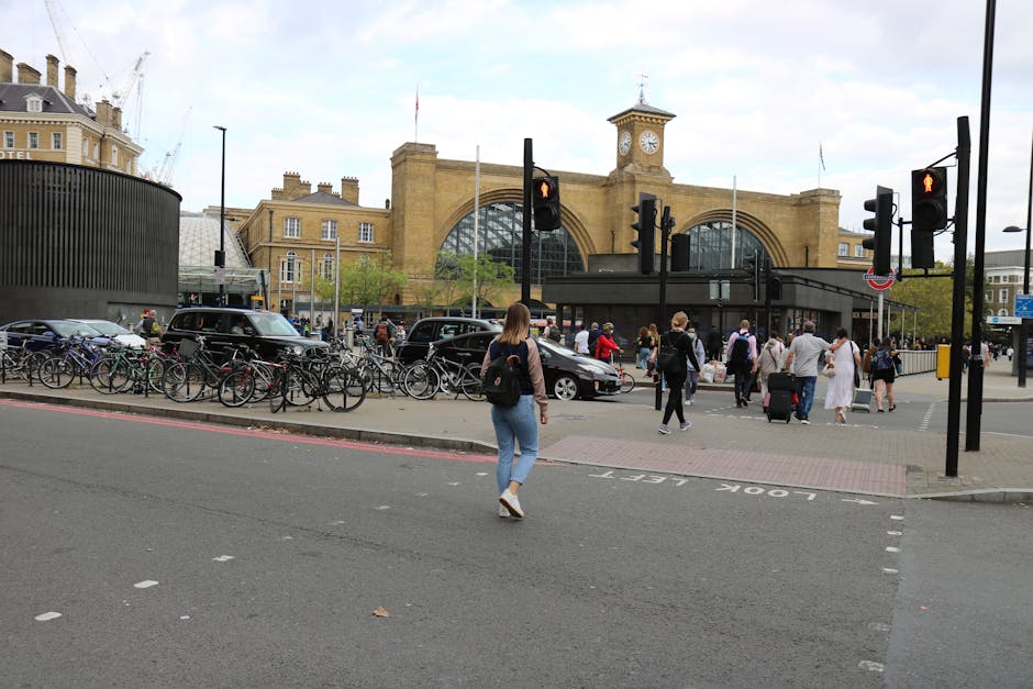 A busy scene outside Kings Cross station showing pedestrians crossing the road amidst traffic lights and vehicles. In the foreground, a woman with a black backpack, wearing a grey top and blue jeans, walks across the street toward the station entrance. To her right, several people are waiting or walking near a group of luggage and shopping bags. Bicycles are parked in designated racks along the pavement, which is separated from the roadway by a curb. The station building, made of brick with large arched windows and a clock tower, dominates the background. Visible inside the station entrance are some commuters and staff, as well as outdoor signage. The environment appears to be daylight with a cloudy sky overhead, and the scene captures urban activity associated with transport hubs, relevant for house removals and moving-related logistics, as managed by Man and Van King's Cross.