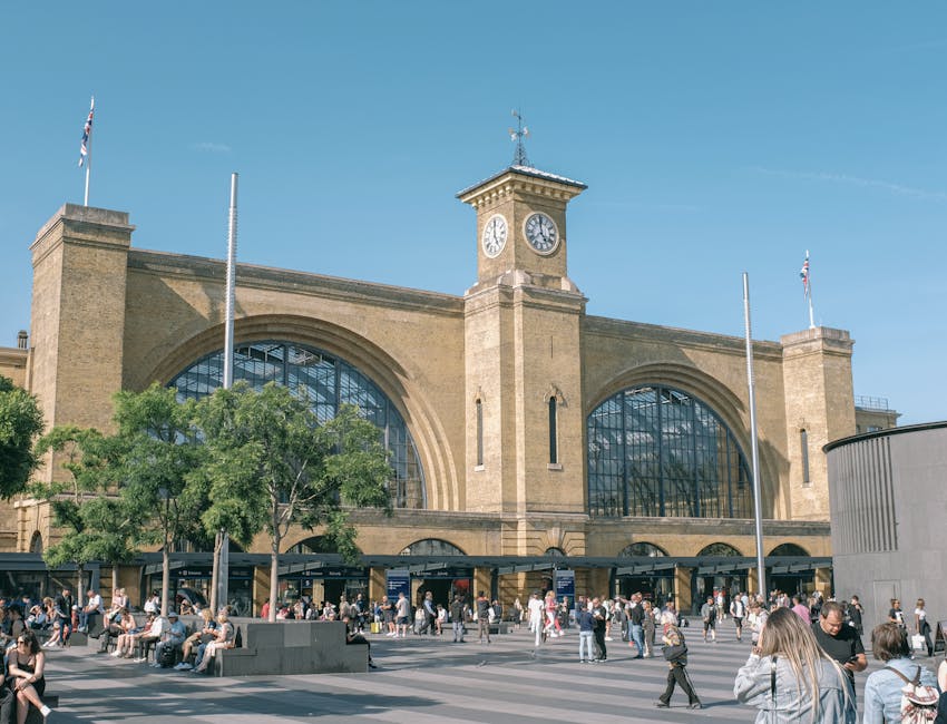 The exterior of Kings Cross railway station in London is depicted with a large, historic brick façade featuring arched windows and a central clock tower with a round clock face, set against a bright blue sky. The station entrance is busy with pedestrians walking on the pavement, some sitting on benches, while others are entering or leaving the station. In front of the building, there are several flagpoles with flags flying. The foreground shows a mix of individuals with luggage, and some are engaged in activities related to home relocation or furniture transport. Green trees partially obscure parts of the station, and the overall scene captures a lively transportation hub often involved in packing and moving logistics, aligning with professional removals services like those offered by Man and Van King's Cross.