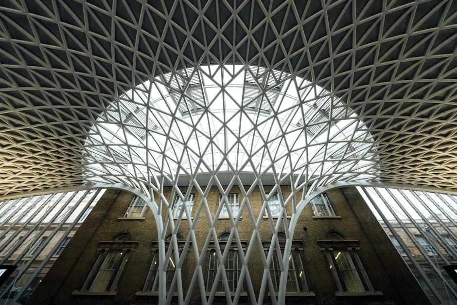 The image shows an interior view of a modern building's central atrium with a distinctive architectural ceiling structure made of white, lattice-like metal beams forming a large, rounded opening in the ceiling. Beneath the ceiling, there is a brick facade with classical-style windows, contrasting with the contemporary roof design. The building is illuminated by natural light filtering through the open lattice, which also reveals external staircases and additional architectural elements visible through the ceiling opening. The setting appears to be within a professional or commercial environment, where the structural details suggest a space that could be associated with office or public buildings suitable for house removals, packing, and loading processes. This visual could support content about transport logistics, furniture transport, and the organisation of home relocation services inside a commercial property, relevant to the loading bay rules at Kings Cross as detailed on the page for Man and Van King's Cross.