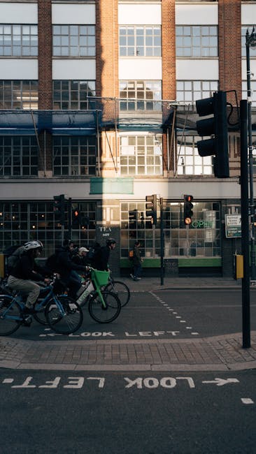 The image depicts a busy urban street scene in front of a multi-storey brick building with large glass windows, which appears to be a commercial or residential property. In the foreground, several cyclists are riding along the road, wearing helmets and casual clothing, while pedestrians walk nearby. A prominent traffic light is positioned on the right side of the image, and a sign indicating a one-way street is visible on the pavement, which is marked with arrows and the words 'LOOK LEFT.' The street is lined with black metal fencing and a small awning above the ground-floor windows. The scene is illuminated by natural daylight, with shadows cast on the sidewalk and road. The environment suggests typical city activity, with ongoing movement and transportation, aligning with the context of a relocation or home removal process that a company like Man and Van King's Cross facilitates, especially around area restrictions and logistics at loading bays and street parking zones.