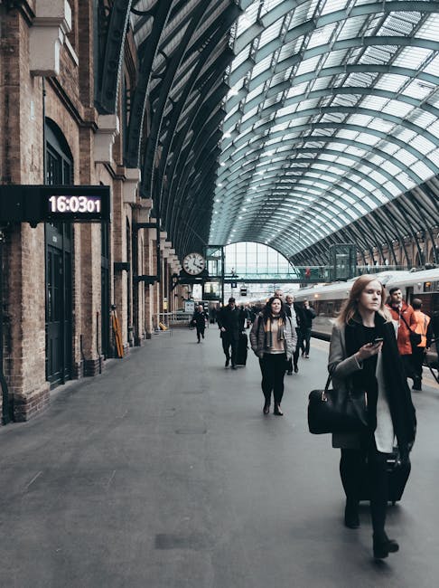 Inside the spacious train station at Kings Cross, there is a high, arched glass and steel ceiling allowing natural light to illuminate the platform. The platform features a large digital clock displaying the time as 16:03:01 and a traditional round clock on the wall background. Several passengers are walking along the platform, carrying personal belongings such as backpacks, handbags, and suitcases. A woman in the foreground is wearing a grey coat, black trousers, and is holding a smartphone in one hand while pulling a dark suitcase with the other. The platform surface is concrete, and there are various luggage trolleys and bags scattered nearby. In the background, a train is visible on the tracks, and station staff or other passengers are positioned near the platform edges. The environment appears busy but orderly, reflecting typical home relocation or moving logistics tasks often coordinated by removals services like Man and Van King's Cross to facilitate furniture transport and packing during house moves.