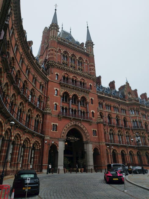 A tall, historic red-brick building with multiple towers and ornate architectural details, including arched windows and decorative stonework, situated in a city centre. The building features a large c