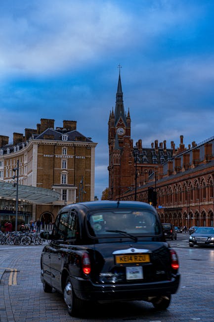 A tall, historic red-brick building with multiple towers and ornate architectural details, including arched windows and decorative stonework, situated in a city centre. The building features a large c